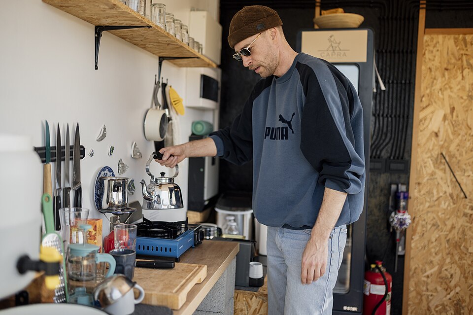 A man making coffee in a small kitchen with open shelves and a knife rail