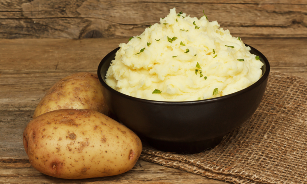 A bowl of creamy mashed potato beside whole potatoes